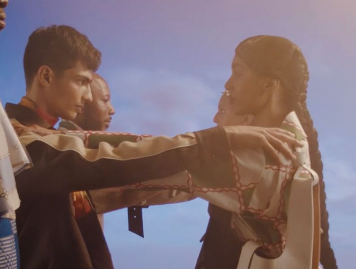 People in clothing face one another with arms extended near the sea, creating a dramatic, solemn scene under a dusky sky.
