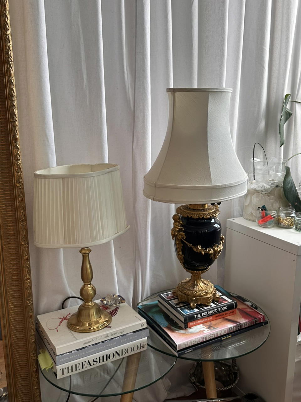 Eclectic, elegant corner with two ornate table lamps, stacked art books on a glass table, gilded mirror, white curtains, and plants.