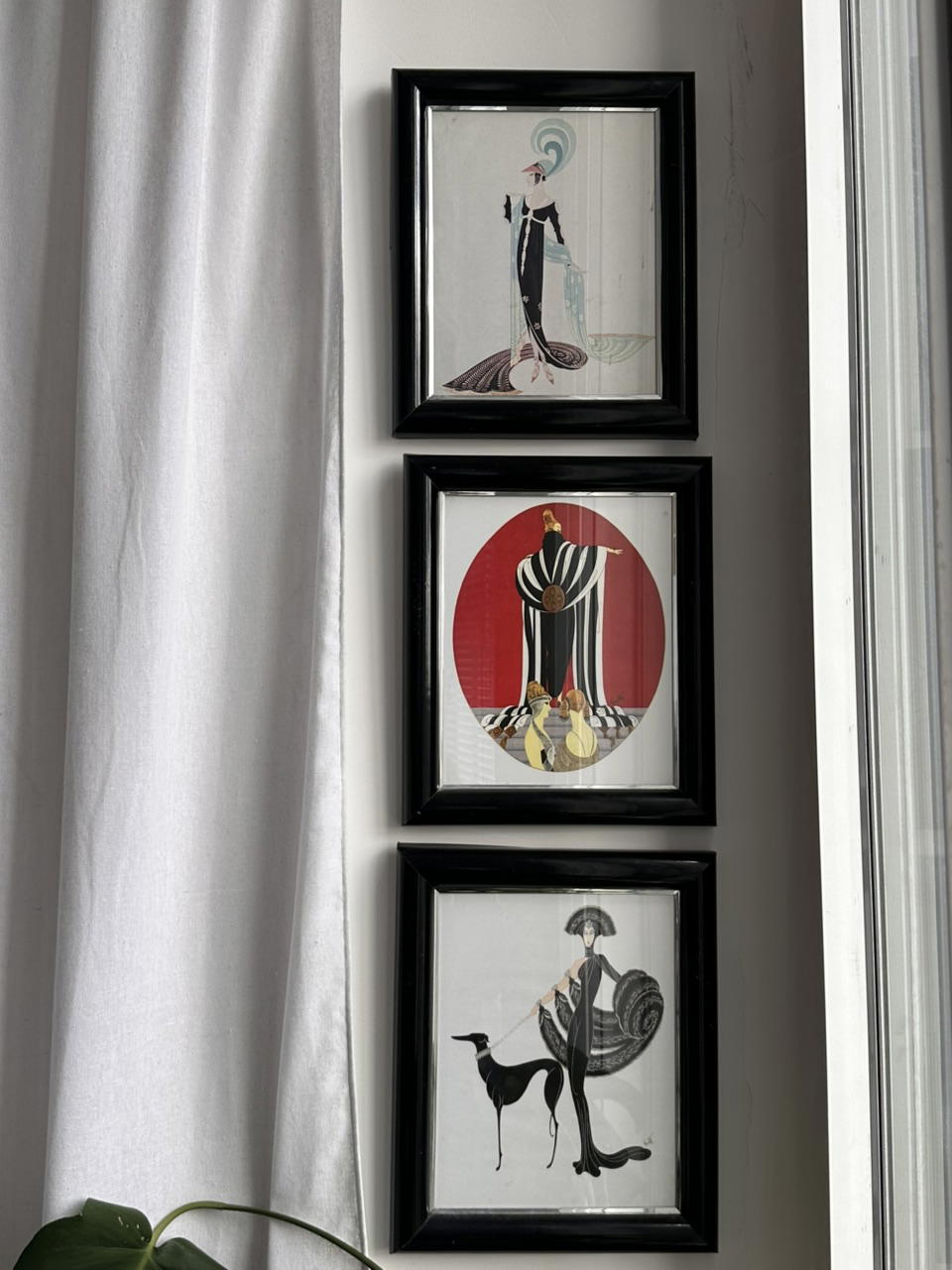 Three black-framed Art Deco fashion illustrations hang vertically beside a bright window and white curtain, creating an elegant vintage display.
