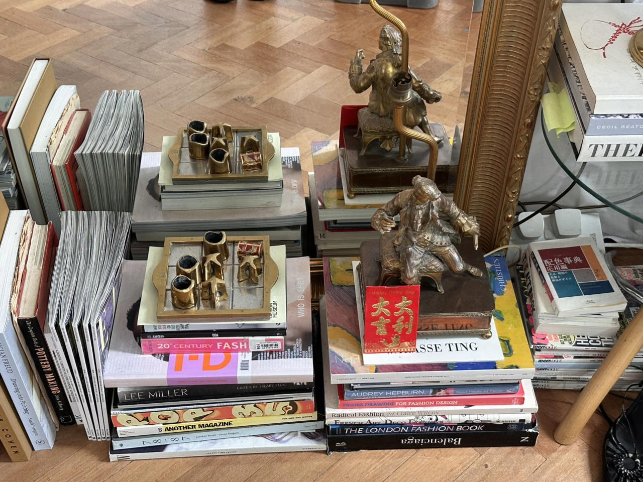 Cluttered bookstore display piled with fashion books and magazines, brass tea sets, bronze figures, and a bright red Chinese ornament.