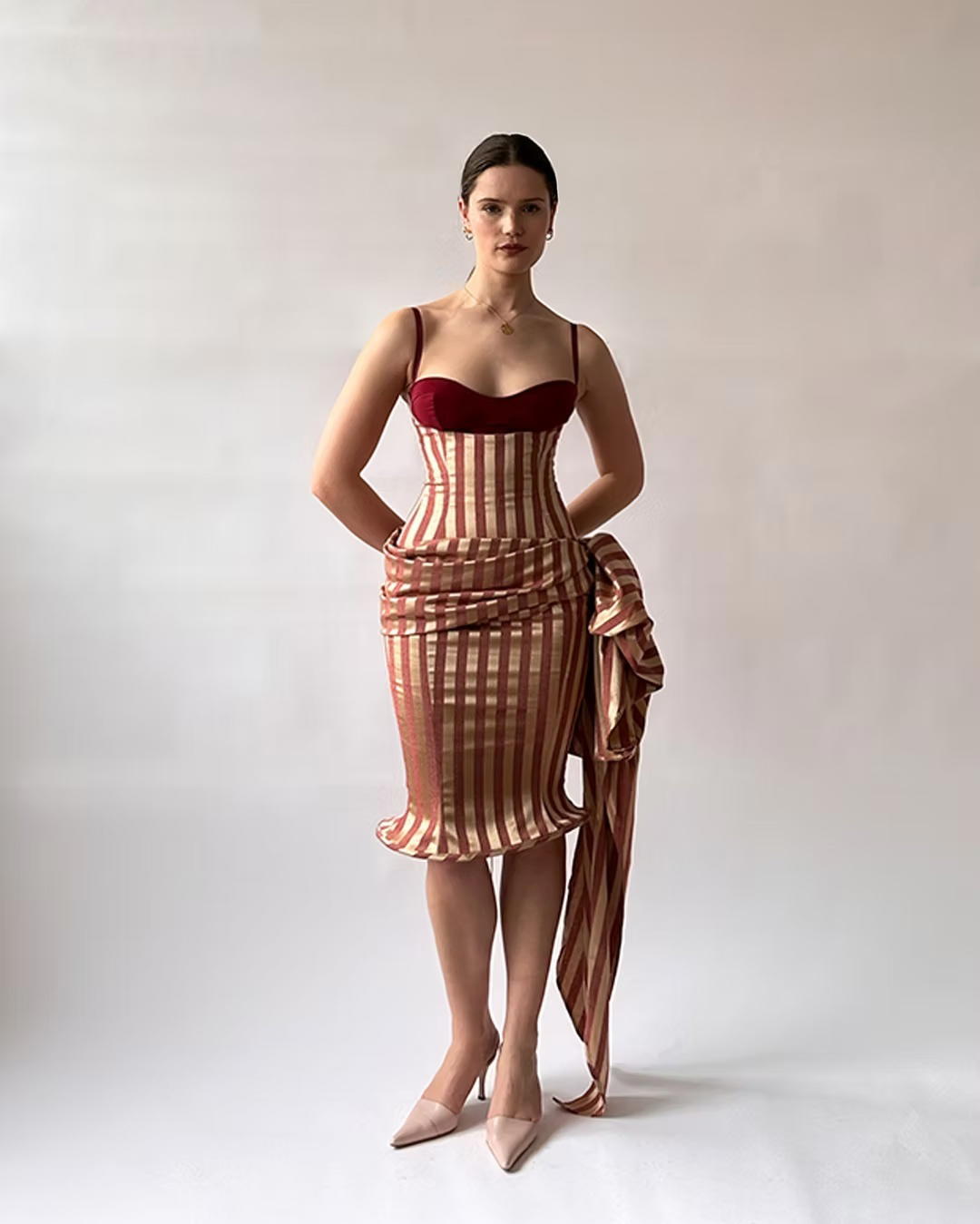 Fashion model poses confidently in a vintage-inspired burgundy and striped corset dress, holding draped fabric against a softly lit studio backdrop.