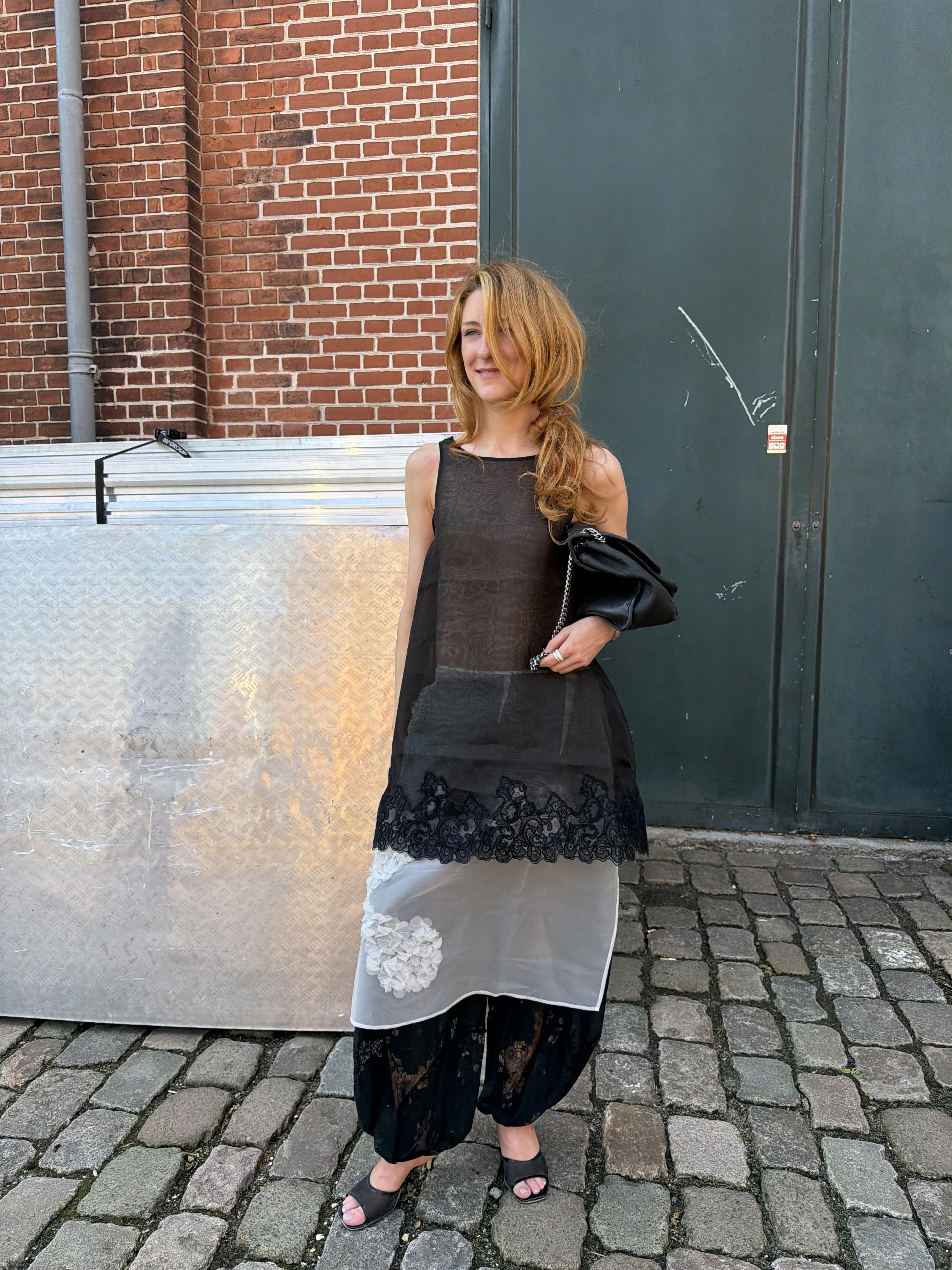 Young woman in a layered black-and-white dress stands on a cobblestone street by a brick wall and dark door, carrying a black purse.