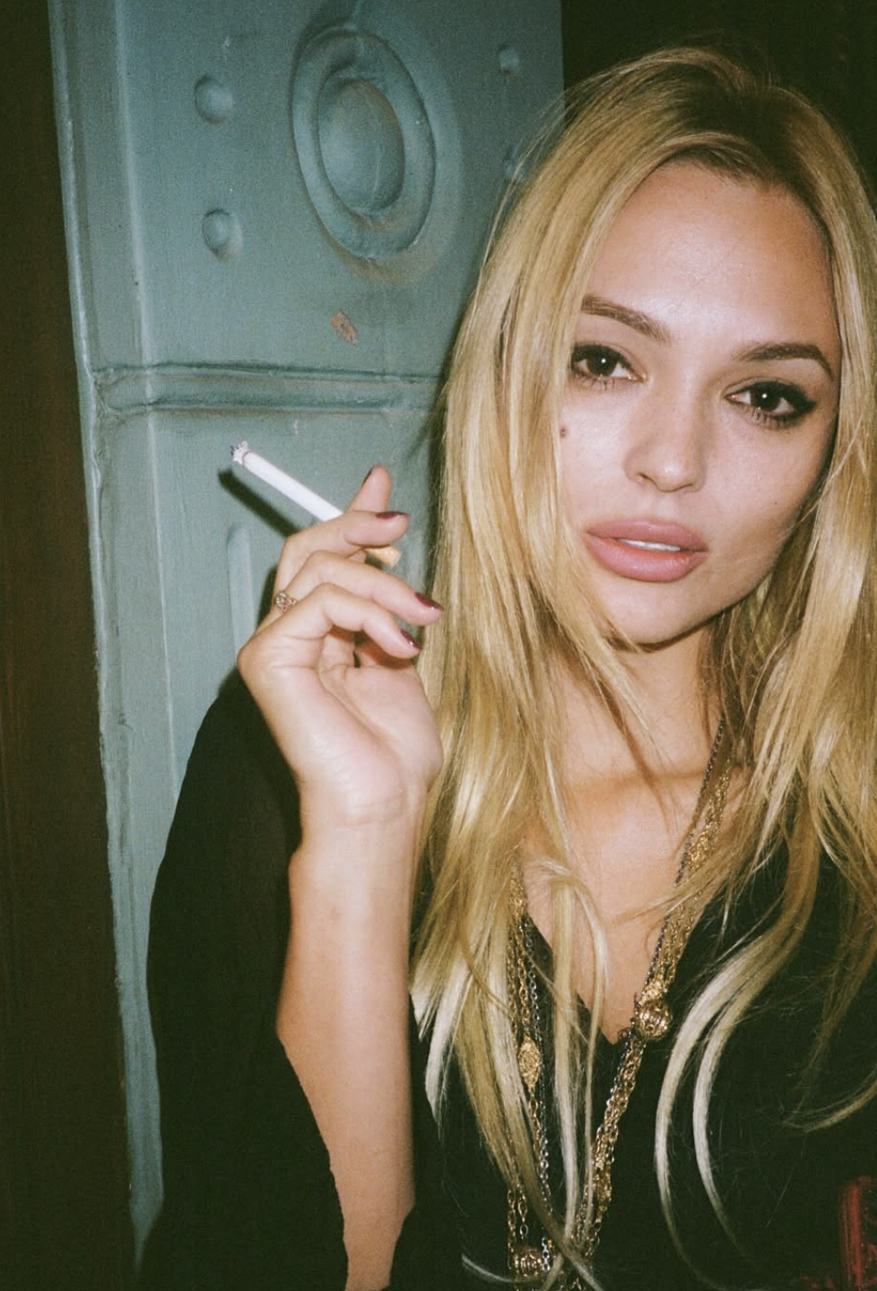 Close-up portrait of a blonde woman holding a cigarette before a teal door, gazing at the camera with a sultry, moody vintage tone.