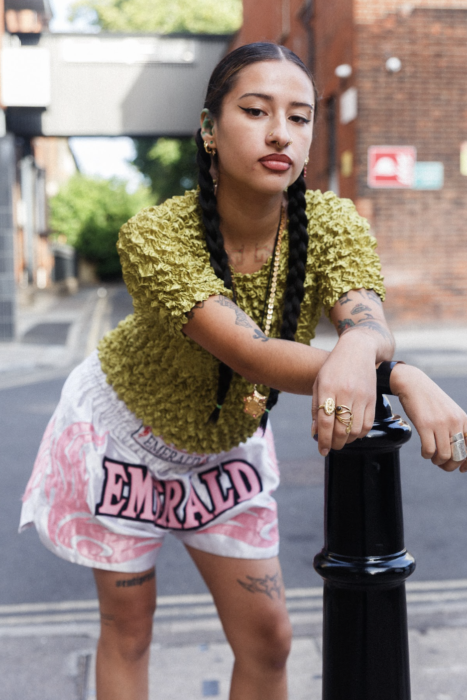 Tattooed woman with braided hair leans on a black street bollard, wearing a textured green top and bold pink-and-white “EMERALD” shorts.