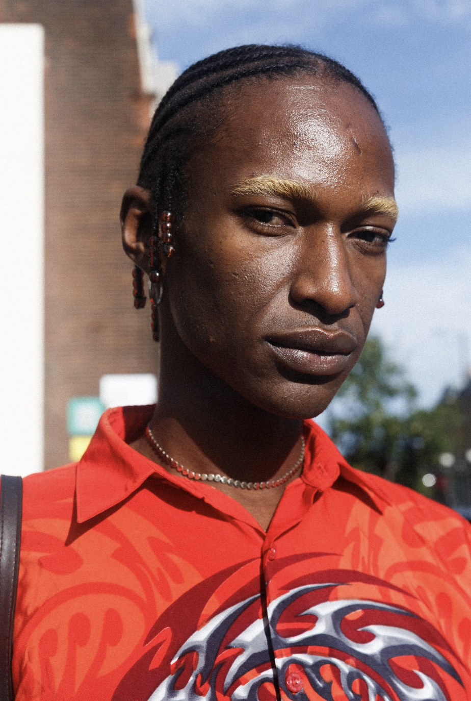 Close-up portrait of a man with braided hair wearing a bright red patterned shirt outdoors, gazing calmly at the camera in sunlight.