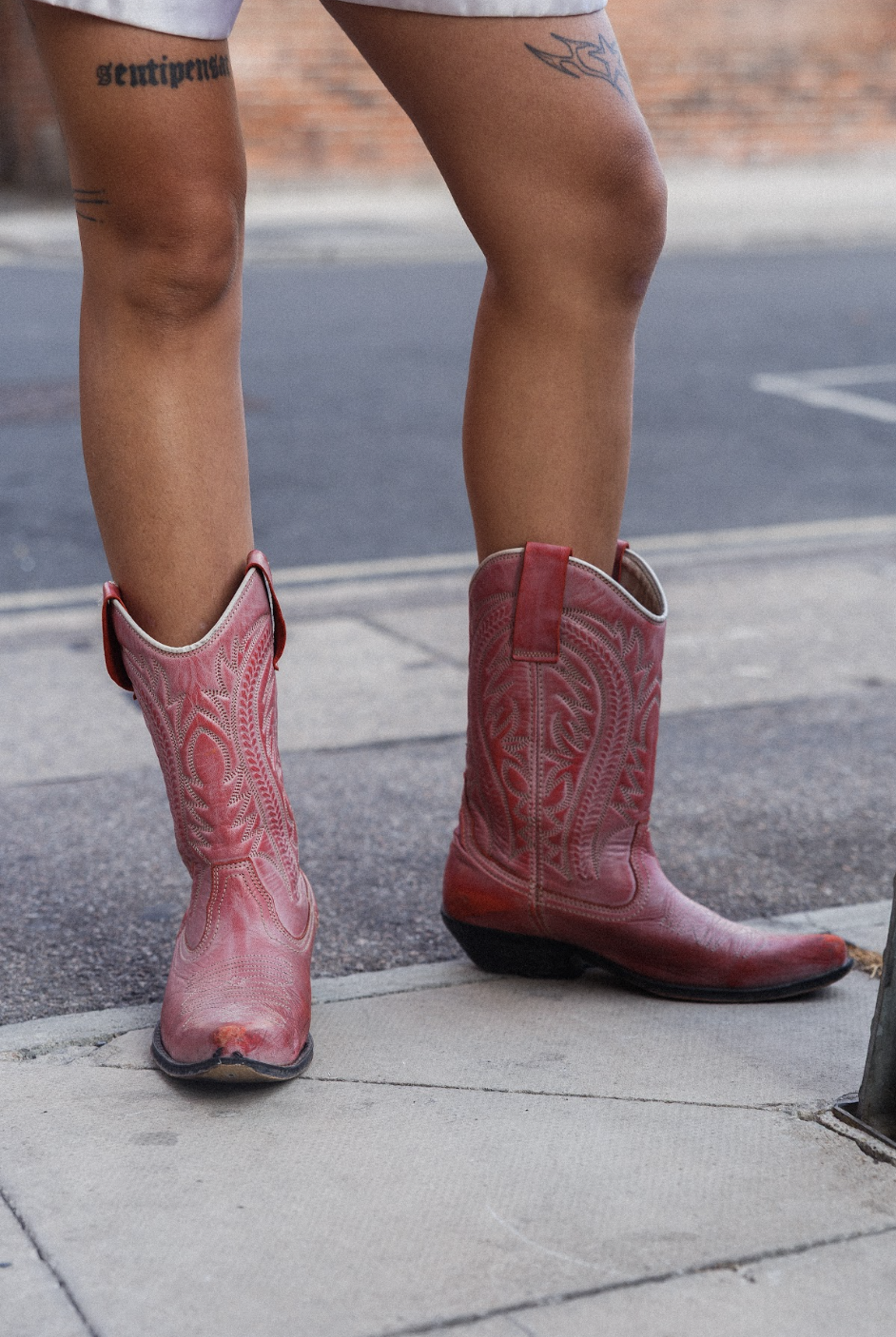 Close-up of tattooed legs in ornate red cowboy boots standing on a city sidewalk, conveying a bold, stylish streetwear mood.