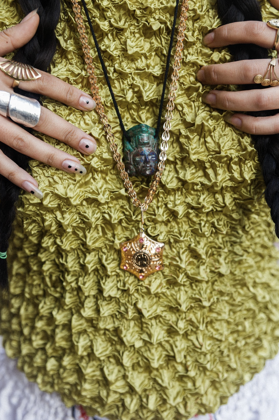 Close-up of hands with rings and nail art resting on a crinkled green garment, layered necklaces and ornate pendants conveying bohemian style.