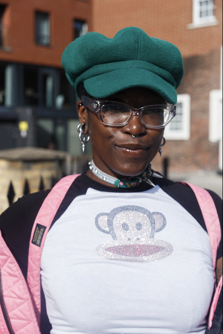 Portrait of a stylish person wearing a teal cap, clear glasses, earrings, pink backpack straps, and a white shirt with a cartoon monkey.
