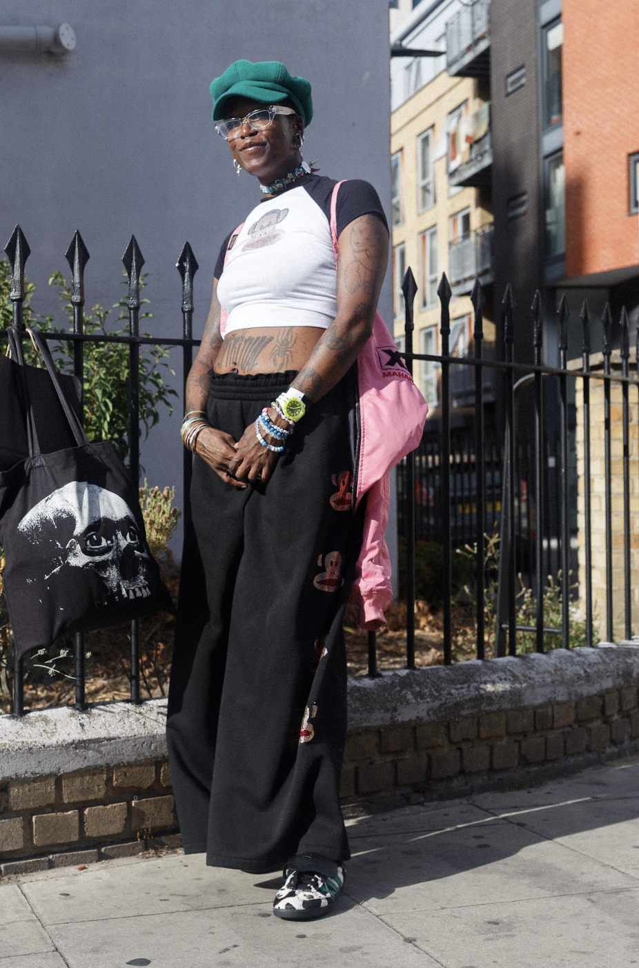 Fashionable person in a green cap, cropped white top, black wide-leg pants, sneakers, and pink bag poses confidently on a city street.