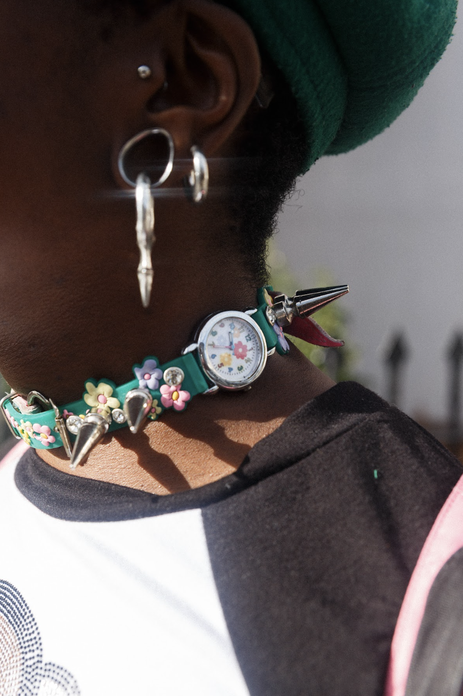 Close-up profile of a person in a green hat, wearing silver earrings and an ornate floral choker with a tiny watch; bold, artistic style.