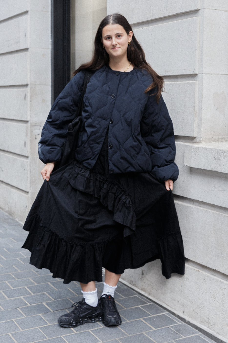Young woman in a black quilted jacket and layered ruffle skirt poses outside a stone building, wearing white socks and black shoes.