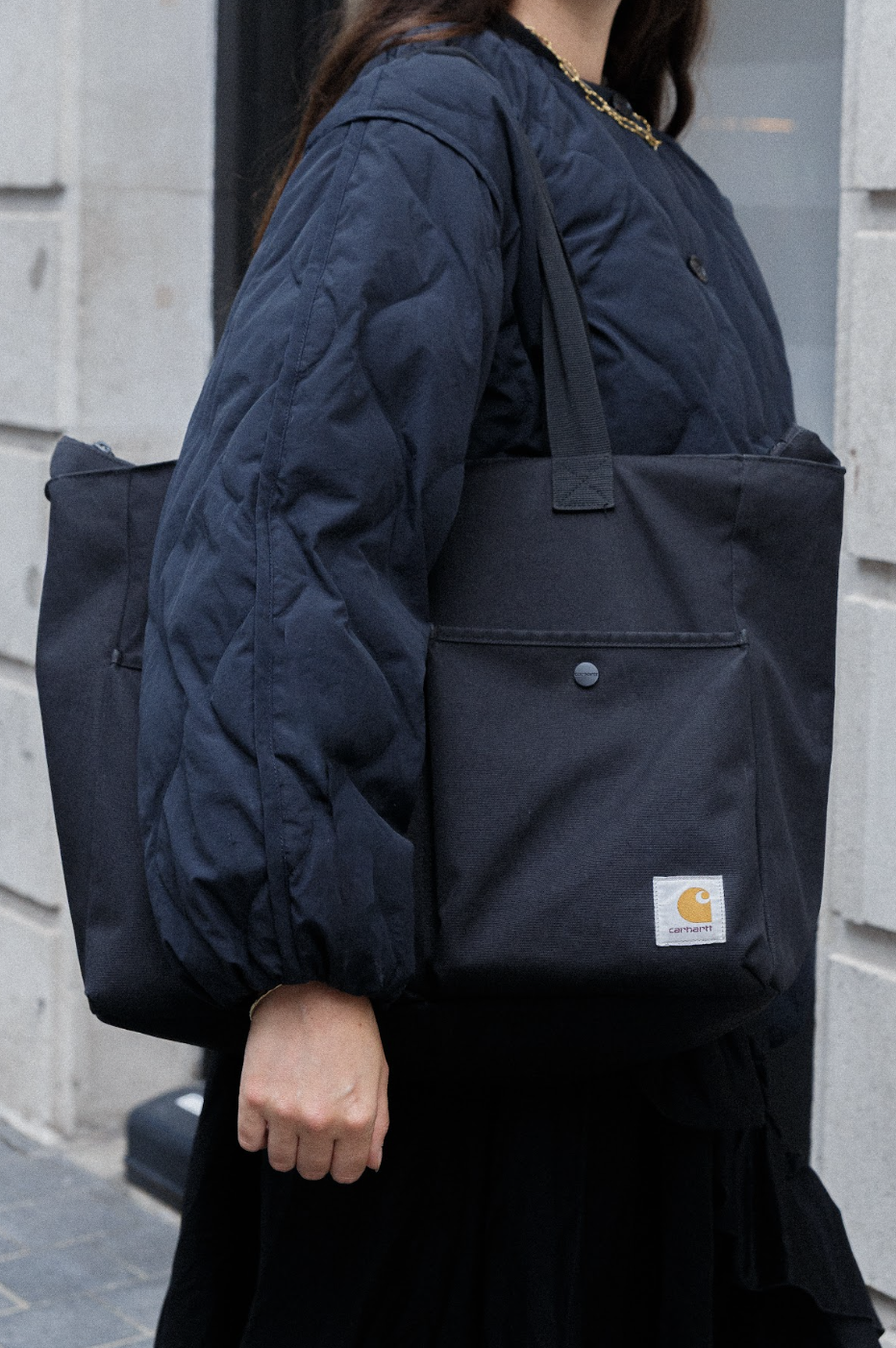 A person in a navy quilted jacket carries a large black Carhartt tote bag against a gray urban wall, conveying a practical, stylish mood.