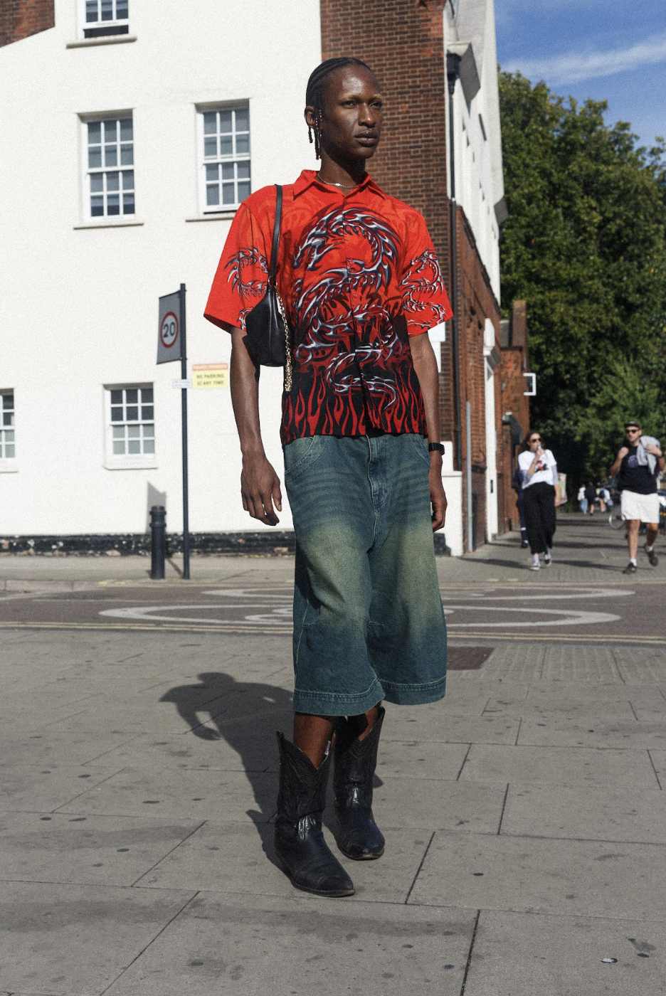 Stylish man in a red graphic shirt, denim shorts, and black boots walks confidently across a sunny city street, casting a long shadow.
