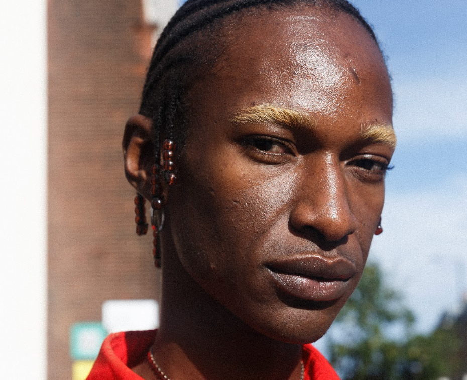 Portrait of a man with braided hair and earrings wearing a vivid red patterned shirt, gazing seriously outdoors in bright sunlight.