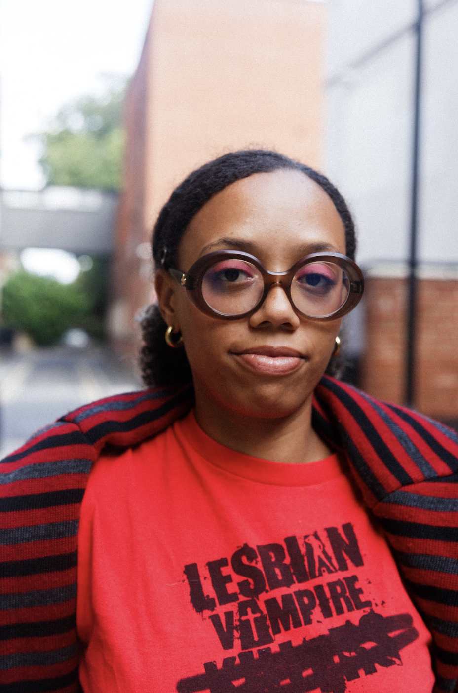 Portrait of a confident woman in round glasses and a red “Lesbian Vampire” shirt, wrapped in a black-and-red striped sweater outdoors.
