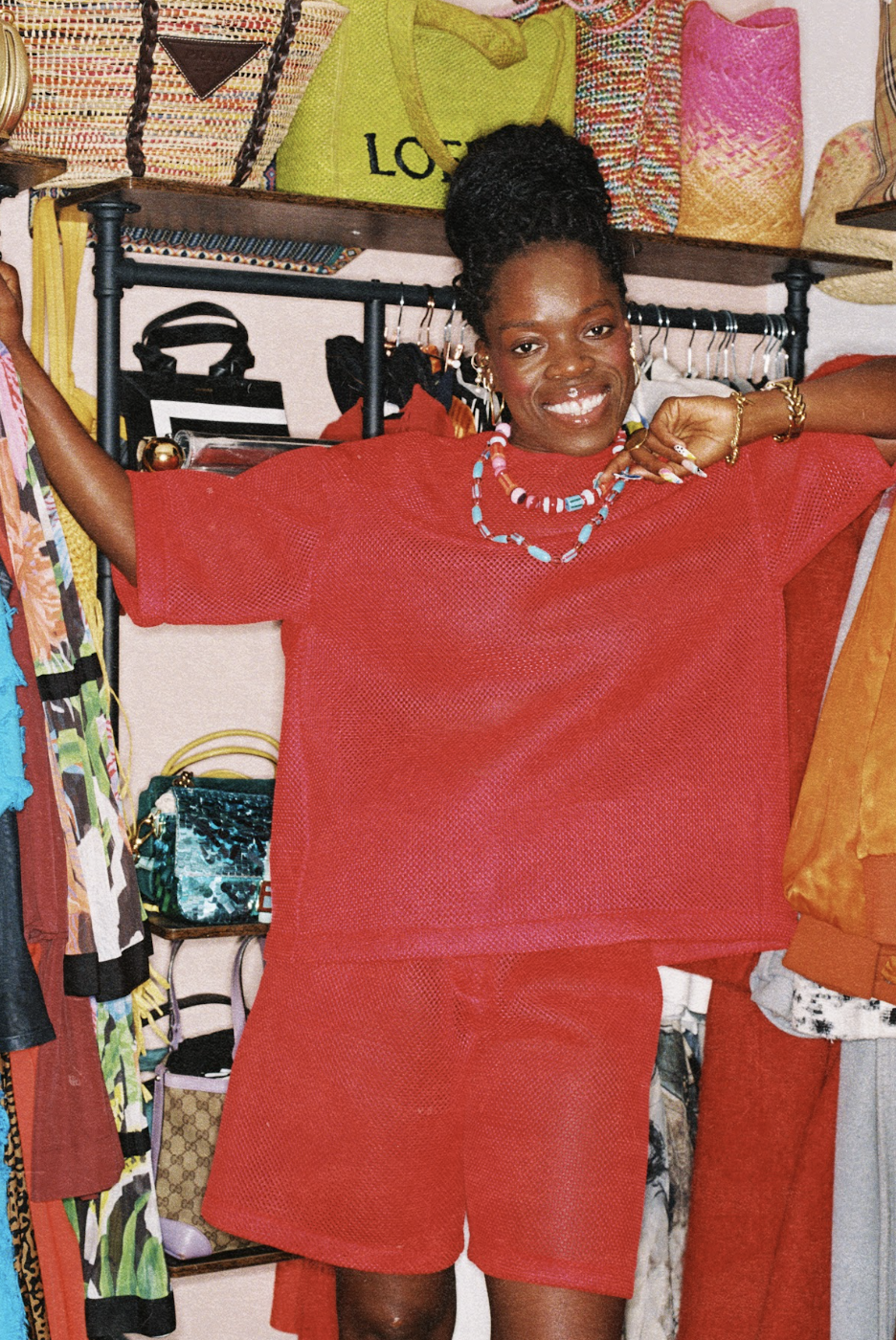 Smiling woman models a bright red two-piece outfit in a colourful wardrobe, surrounded by hanging garments and woven bags.