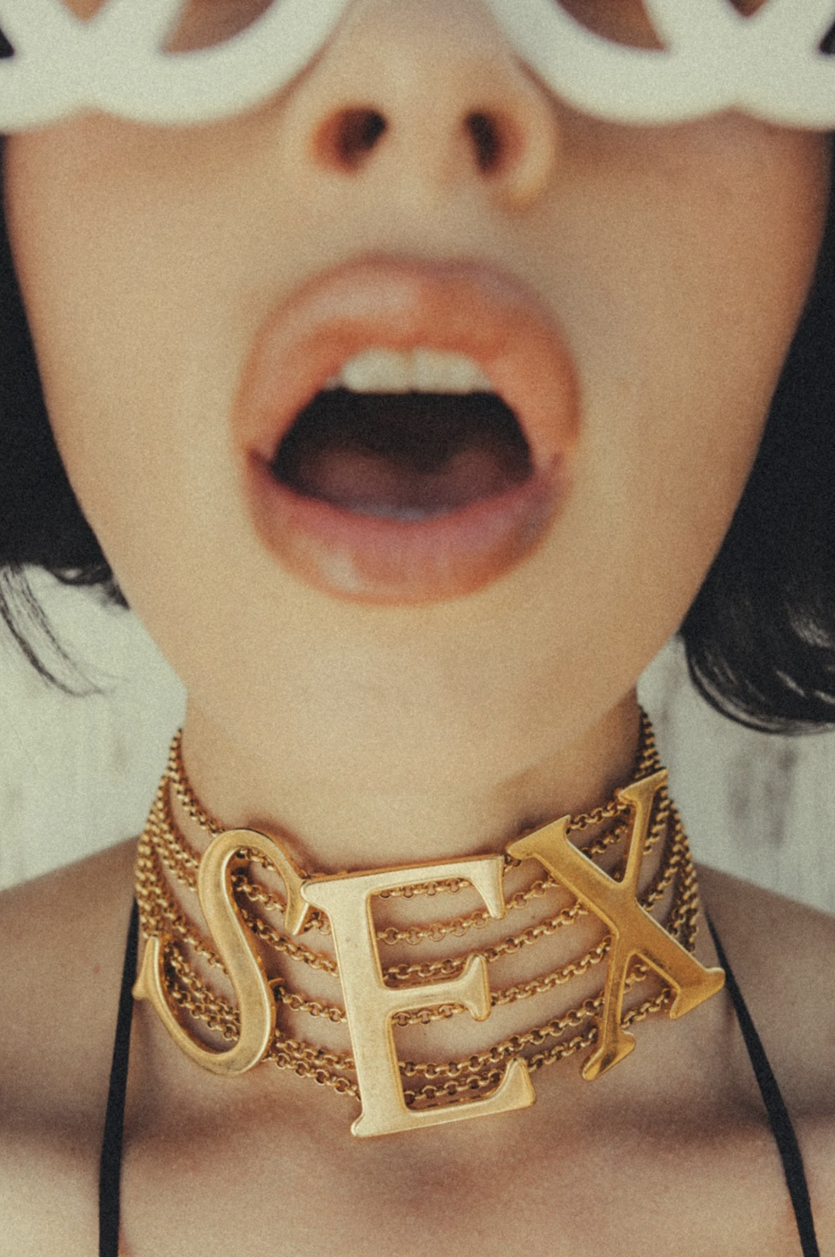 Close-up of a person with an open mouth, white sunglasses, and layered gold chains spelling “SEX,” creating a bold, glamorous fashion mood.