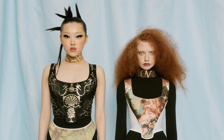 Two fashion models pose in avant-garde black bodysuits, patterned tights, platform heels, and dramatic hairstyles against a pale blue backdrop.