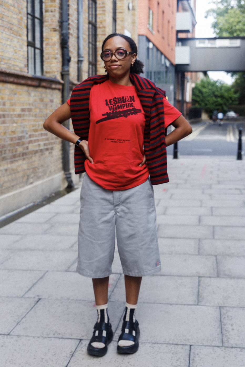 Confident woman in glasses poses hands-on-hips in a red graphic T-shirt and striped cardigan on a city sidewalk, upbeat and stylish.