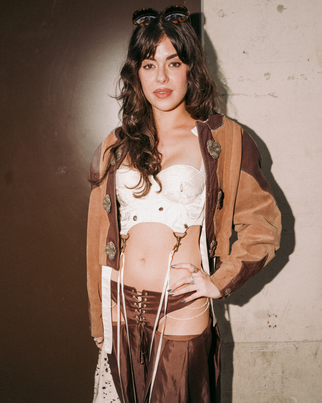 Portrait of a woman with wavy dark hair and sunglasses, posing confidently in a white bustier, brown patchwork suede and leather jacket with vintage hardware and brown leather chaps. Retro, edgy tone.