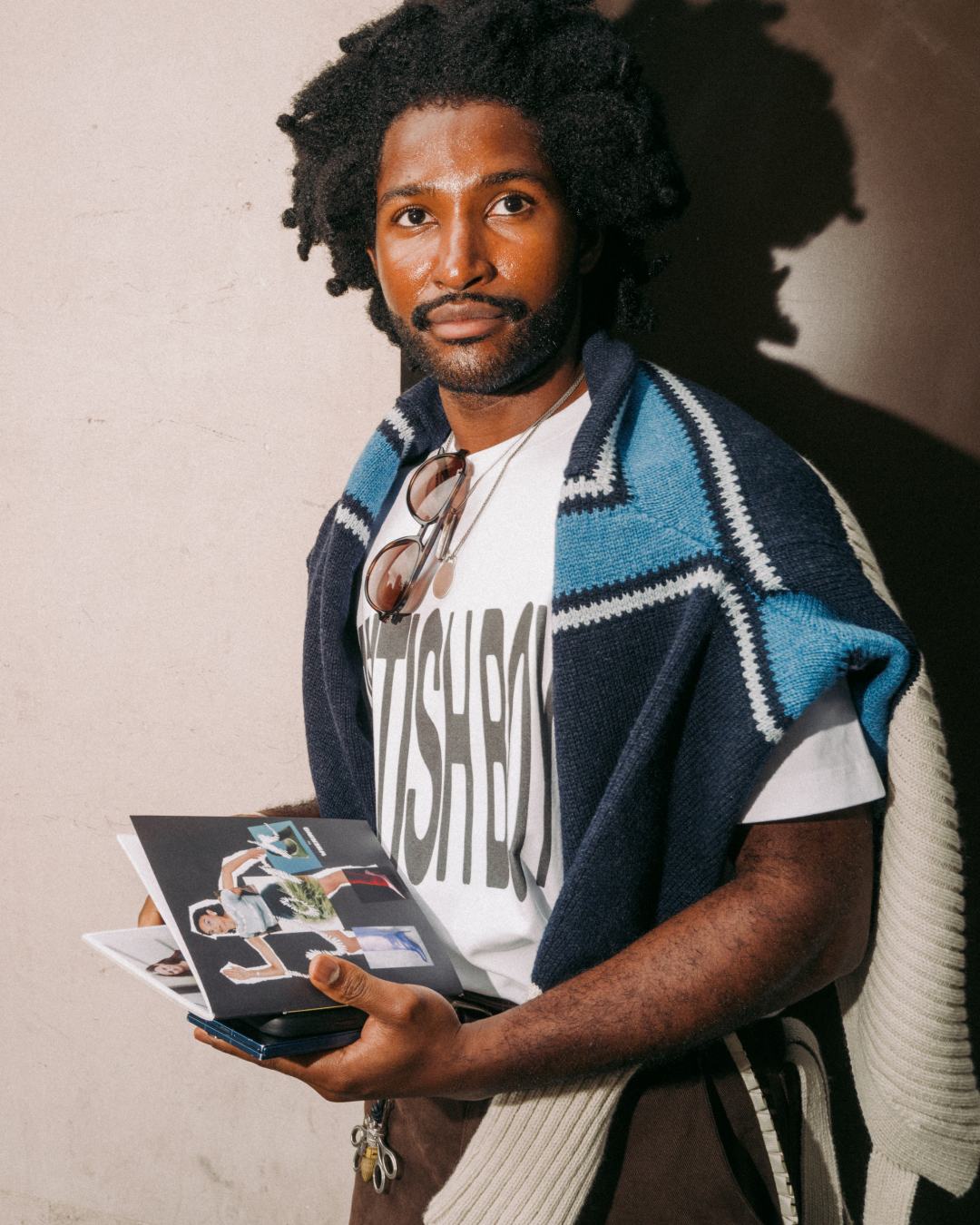 Portrait of a smiling Black man with afro holding an open zine, wearing a blue patterned jumper over shoulders and graphic T-shirt saying "British Boy"; warm, artistic tone.