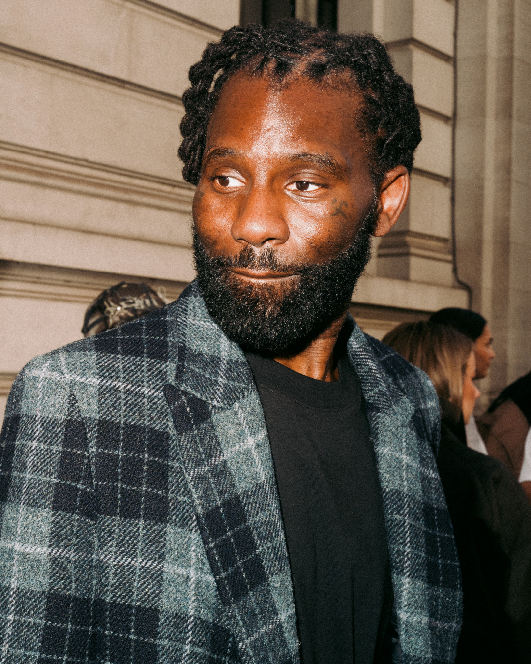 Wretch 32 in a gray plaid blazer and black shirt looks to the side arriving at a fashion show