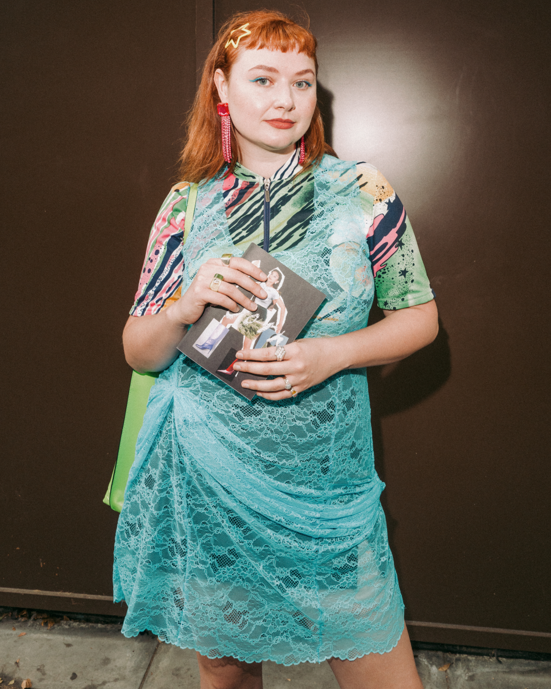 Red-haired woman in a turquoise lace dress and red earrings holds a zine, standing against a dark wall with a poised, confident expression.