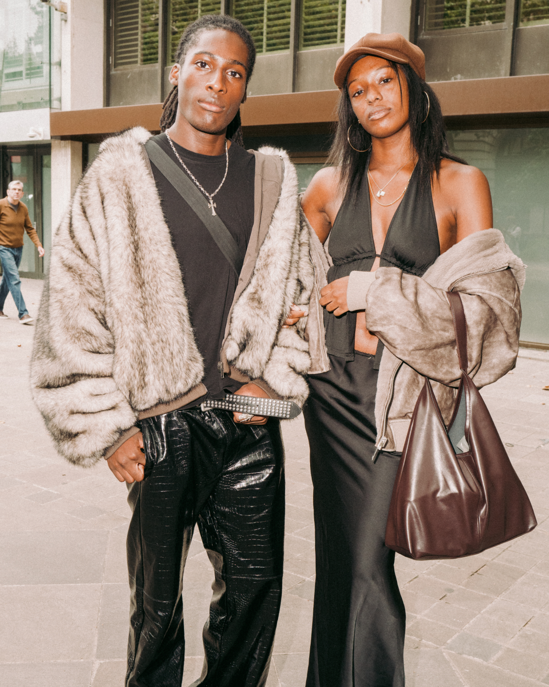 Two stylish Black adults pose outside a city building: one in a fur coat and leather pants, the other in a hat, black dress, and brown bag.