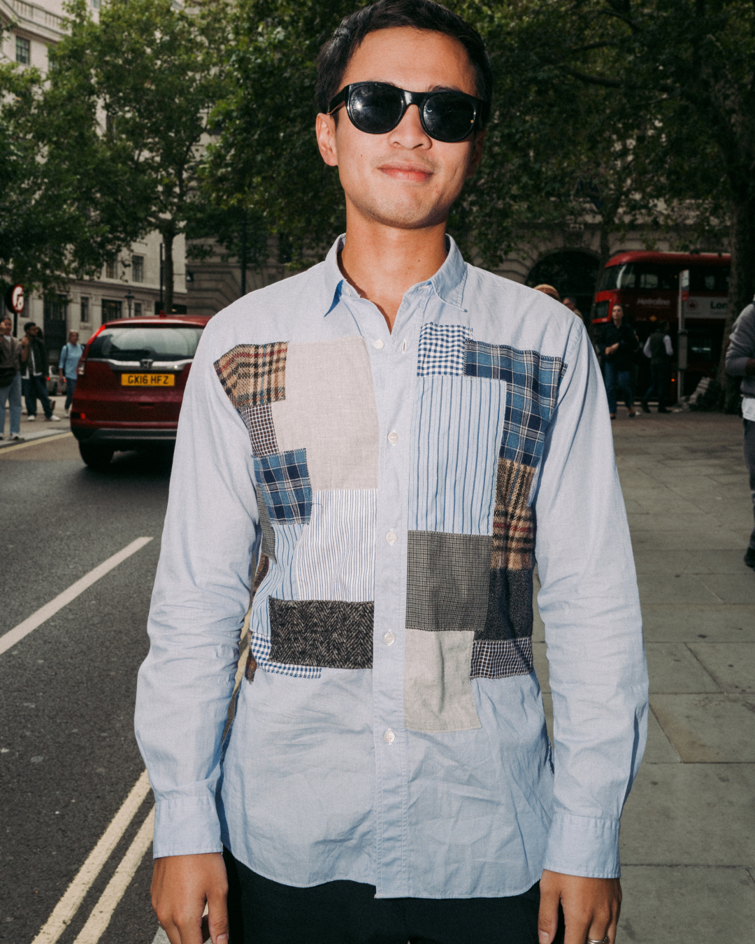 Smiling man in sunglasses wears a light blue patchwork shirt while standing on a city street, with trees, cars, and buses behind him.