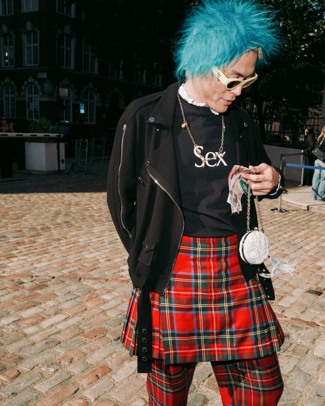 Person with bright blue hair and sunglasses wears a black jacket, “Sex” T-shirt, and red tartan skirt over red tartan trousers on a cobblestone street.