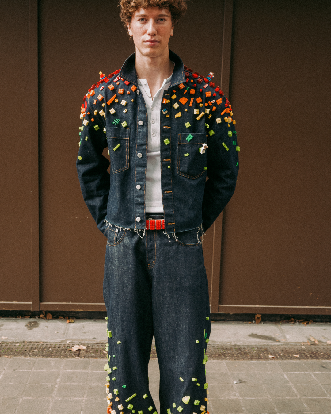 Young man poses confidently outdoors in a denim jacket and wide-leg jeans, colourful lego pieces adding a bold, edgy streetwear style.