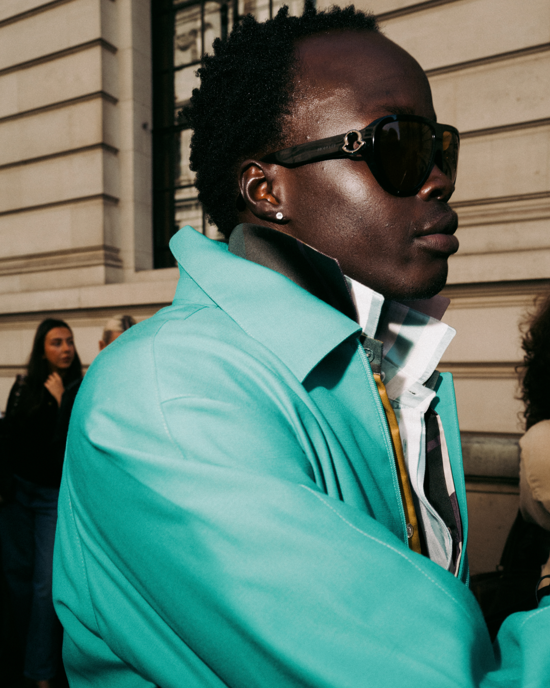 Side-profile portrait of a stylish Black man in sunglasses and a turquoise jacket, standing outdoors by a stone building with a confident, urban mood.