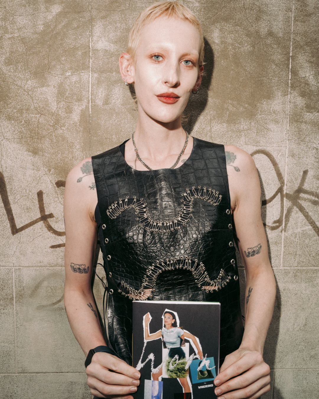 Androgynous person with short blond hair, tattoos, and red lipstick holds a zine, wearing a black studded top against graffiti.