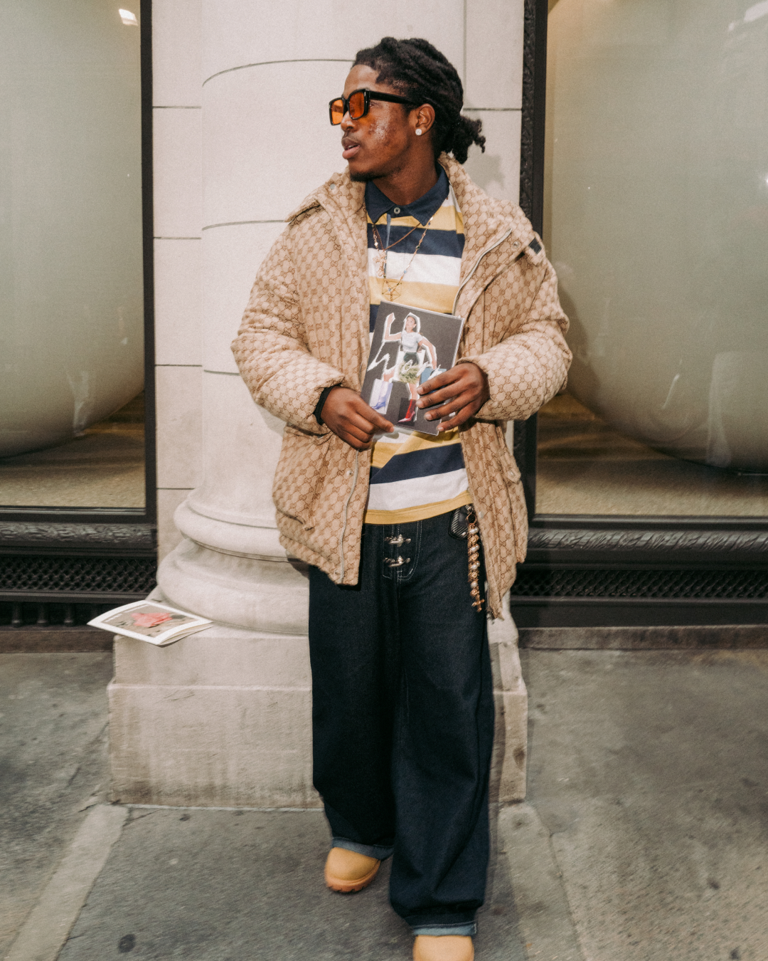 Fashionable man with dreadlocks, sunglasses, and a textured coat holds a magazine outside a stone building, projecting relaxed urban style.