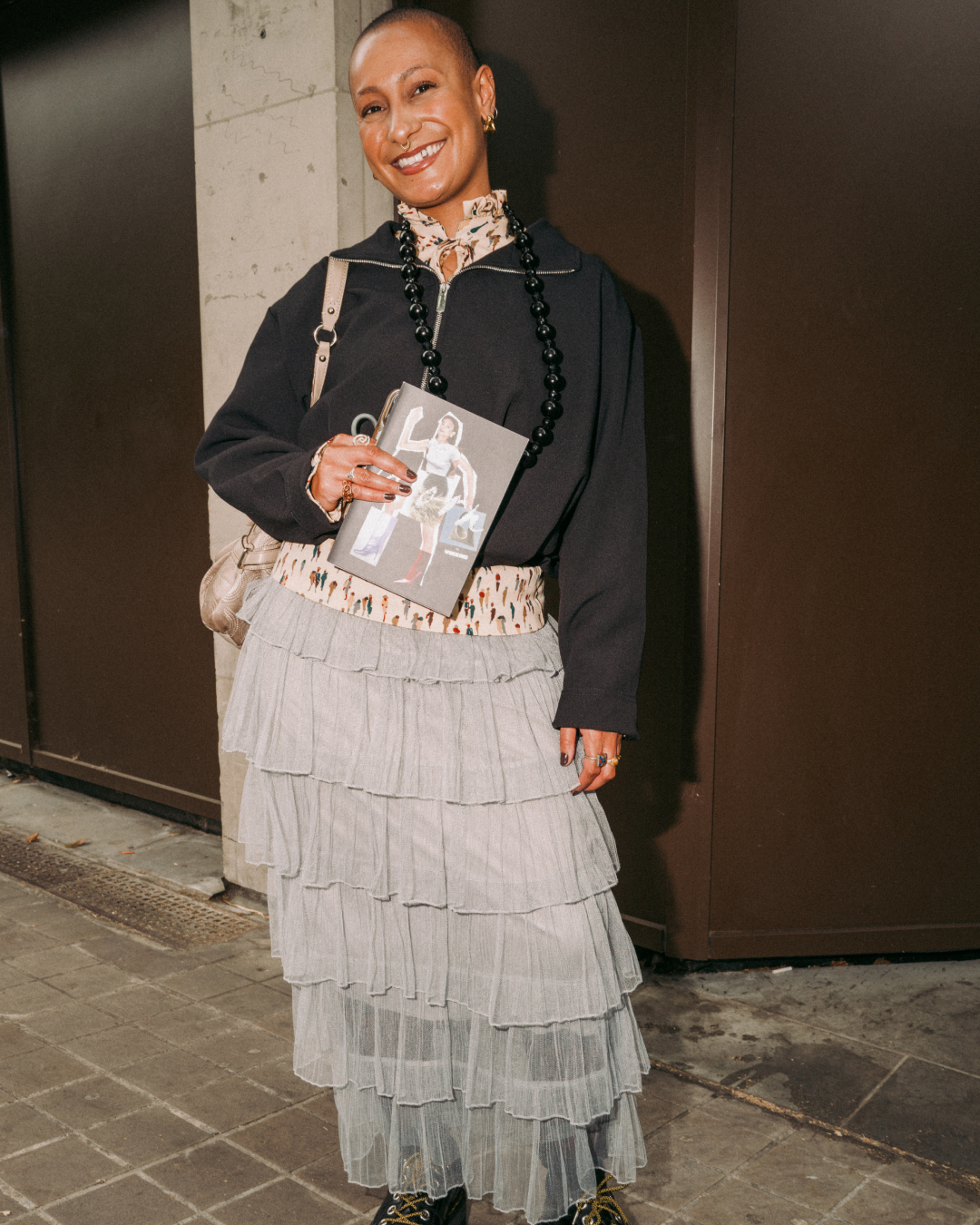 Smiling fashion-forward woman with a shaved head wears a black top and layered gray skirt, holding a magazine