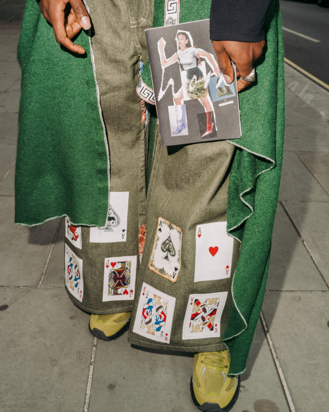 Close-up of a person holding a zine in front of green outfit and trousers decorated with playing cards 