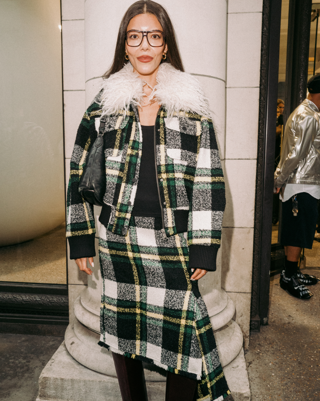 Woman wearing glasses and a green, black, white plaid coat with a feathered collar poses outside a building