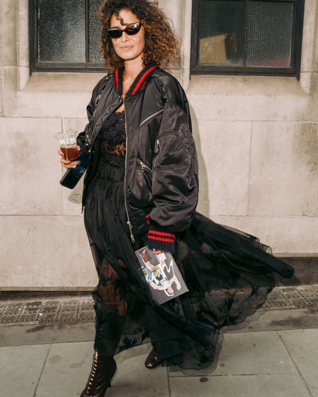 Woman in sunglasses wears a black bomber jacket and flowing dress, holding a drink and magazine on a city sidewalk; confident street style.