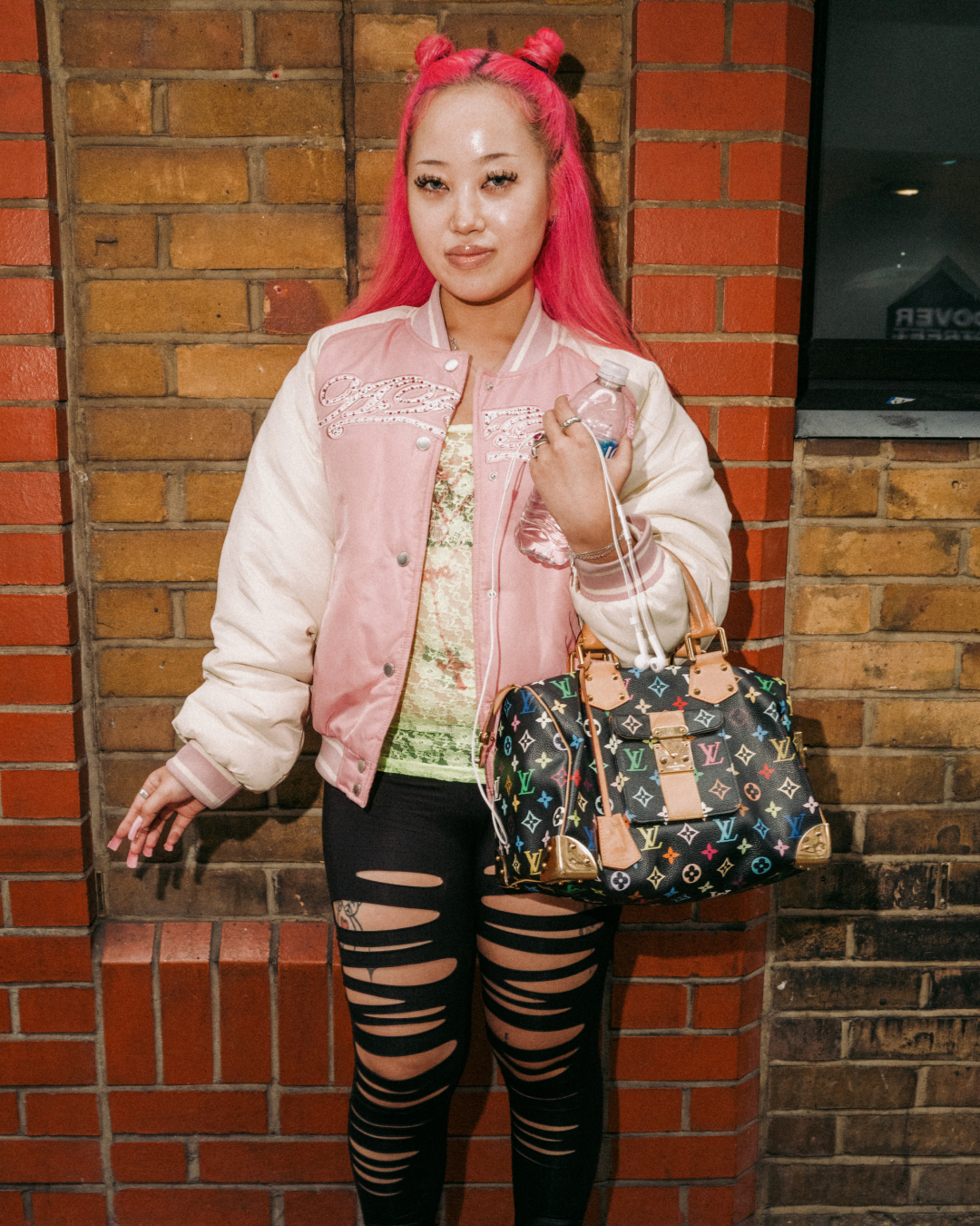 Confident woman with bright pink hair poses against a brick wall, wearing a pink jacket, ripped leggings, and carrying a designer handbag.