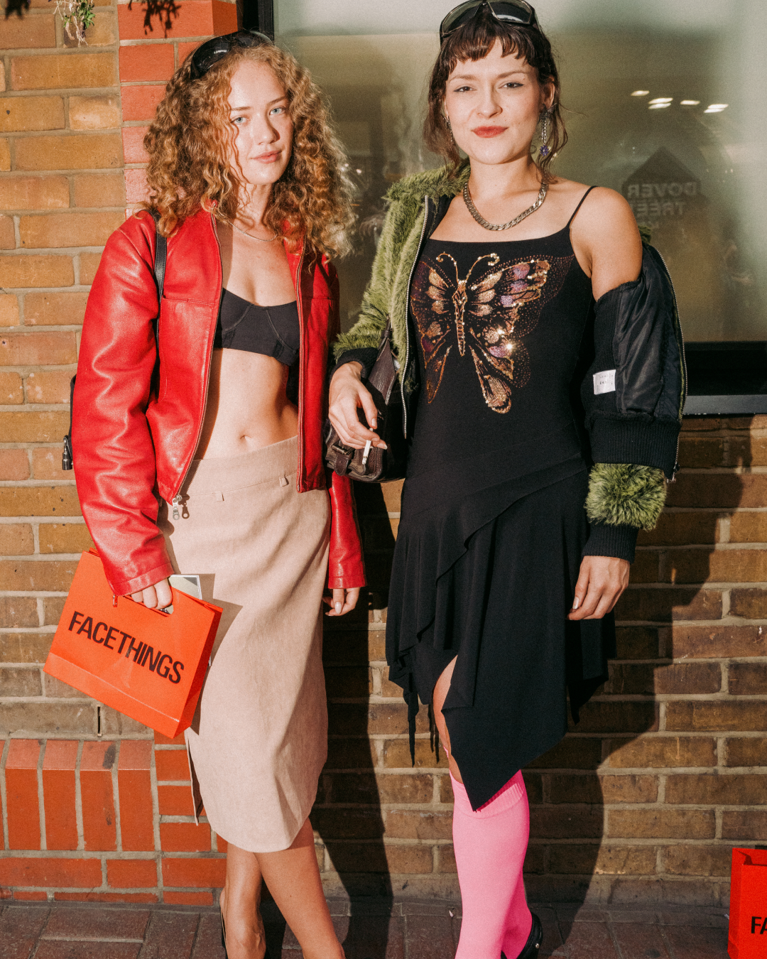 Two stylish women pose outside a brick building; one wears a red leather jacket, the other a butterfly-embellished black dress and pink tights.