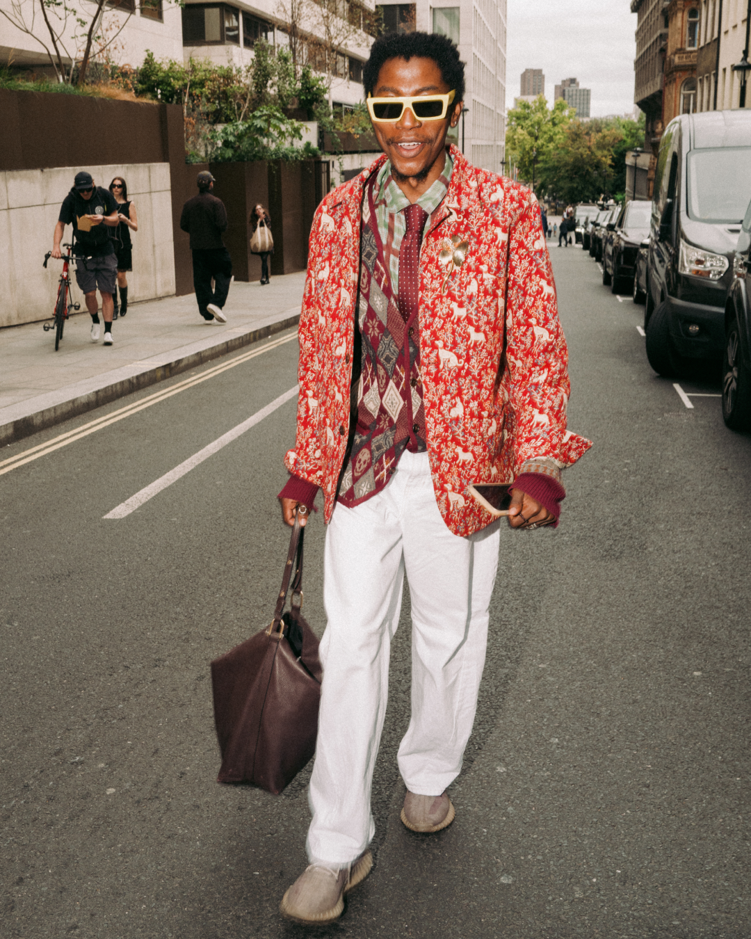 Smiling man in bold red floral jacket, yellow sunglasses, and white trousers walks down an urban street carrying a brown leather bag.