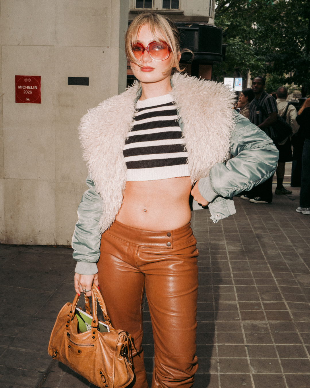 Fashionable woman in red sunglasses, fuzzy jacket, striped crop top and brown leather pants, posing confidently on a city street with handbag.