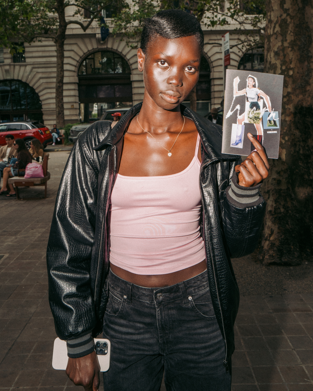 Young Black person in a black leather jacket and pink top poses on a city street, holding a zine